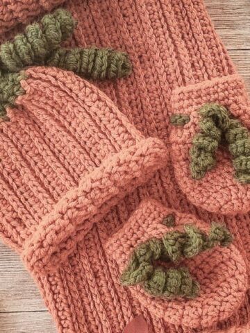 Vintage-style photo of a handmade crochet pumpkin baby set, including a ribbed cocoon sack with button closure, matching hat with green stem detail, and two booties, all styled on a rustic wooden background with scattered autumn leaves.
