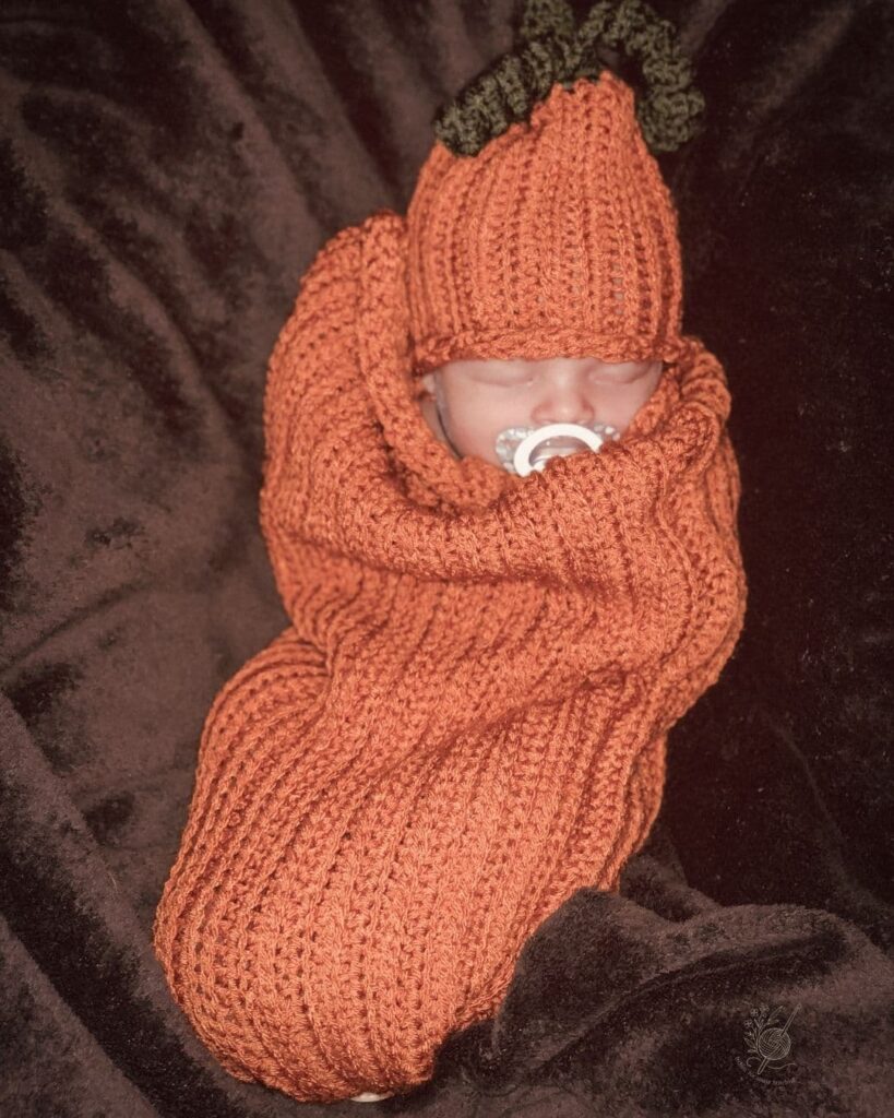 Baby wrapped in a bright orange crochet pumpkin cocoon with matching hat — the perfect fall outfit for newborn photos or Halloween.