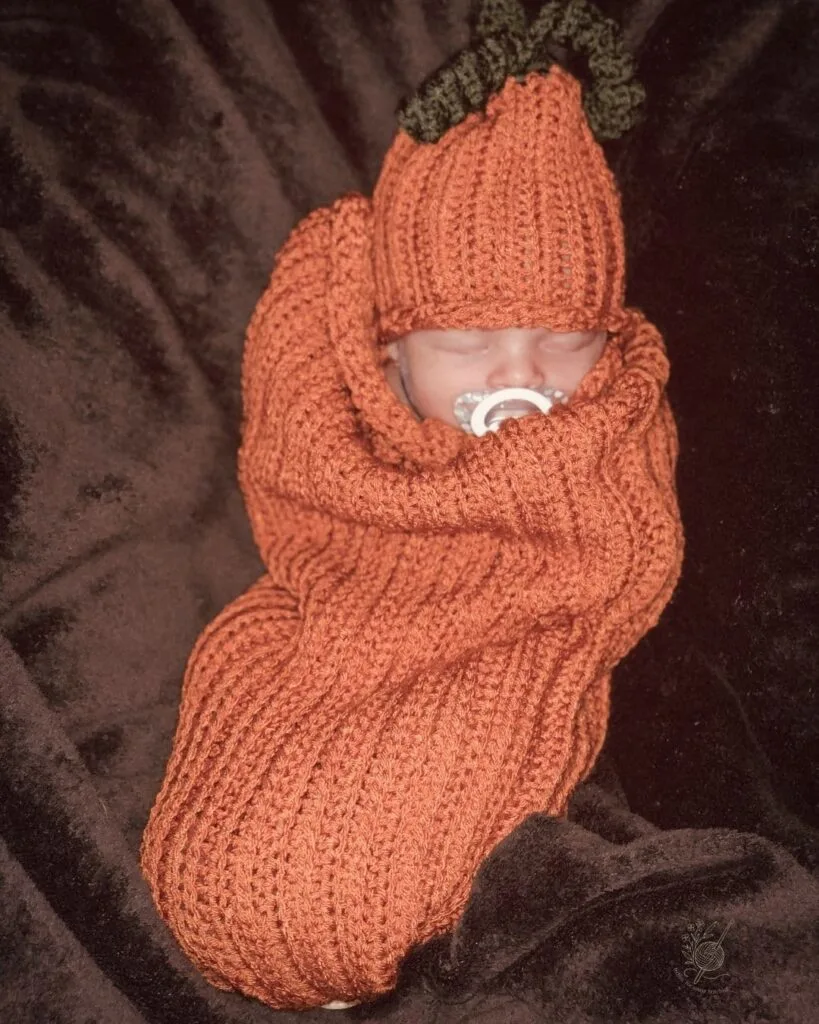 Baby wrapped in a bright orange crochet pumpkin cocoon with matching hat — the perfect fall outfit for newborn photos or Halloween.