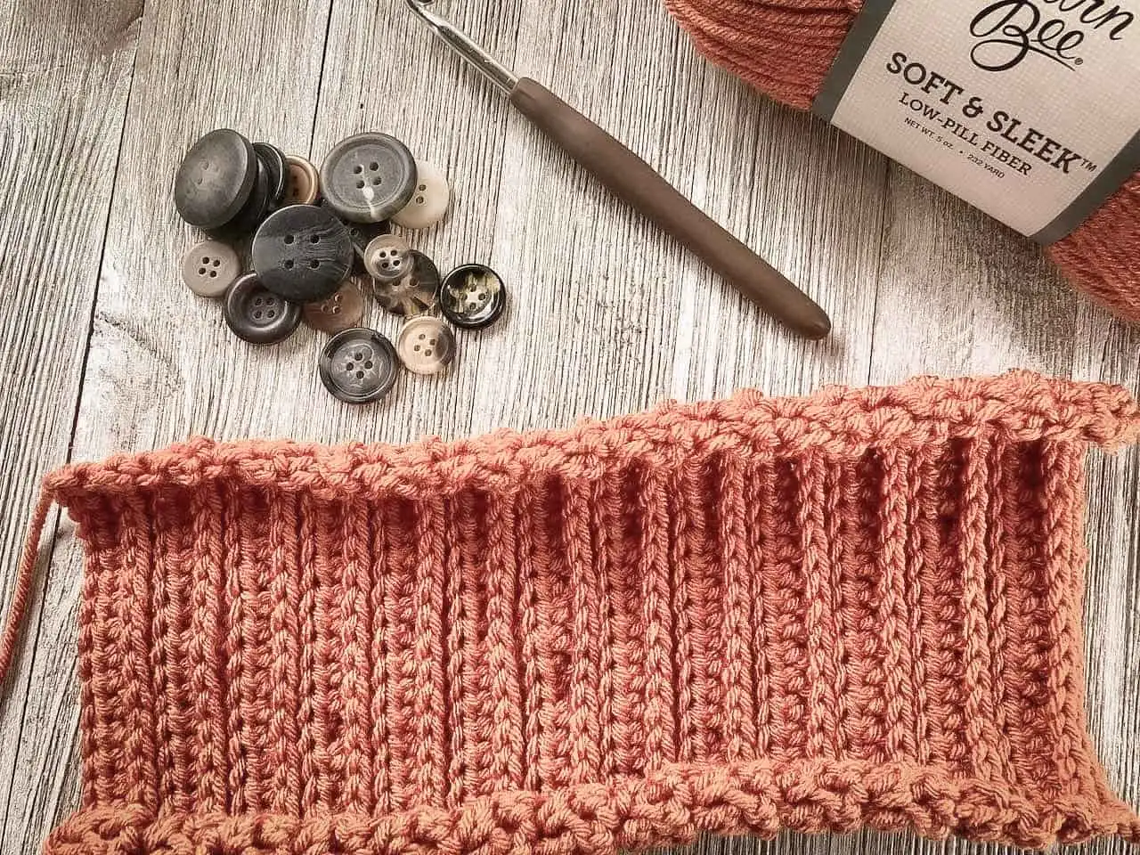 Work-in-progress crochet project in burnt orange yarn, shown with a ribbed stitch swatch on a rustic wood background. Beside the swatch are assorted vintage-style buttons, a crochet hook, and a skein of Soft & Sleek yarn.