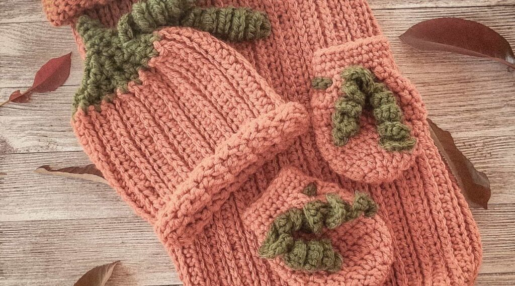 Vintage-style photo of a handmade crochet pumpkin baby set, including a ribbed cocoon sack with button closure, matching hat with green stem detail, and two booties, all styled on a rustic wooden background with scattered autumn leaves.