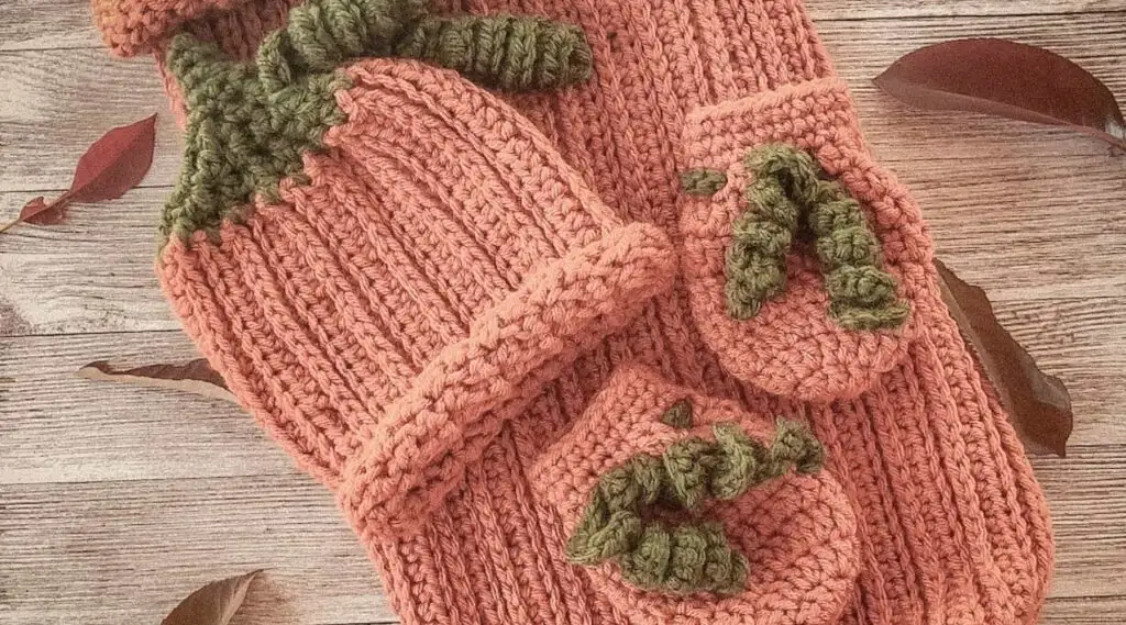 Vintage-style photo of a handmade crochet pumpkin baby set, including a ribbed cocoon sack with button closure, matching hat with green stem detail, and two booties, all styled on a rustic wooden background with scattered autumn leaves.