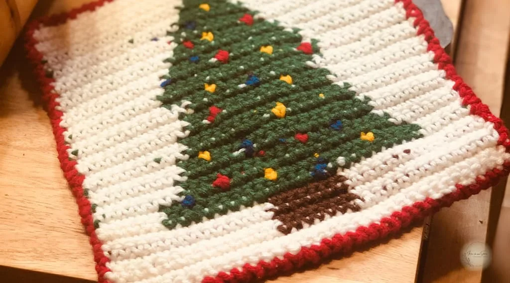 Tapestry crochet Christmas tree potholder with a green tree design, red border, and colorful accents, shown as a free crochet pattern.