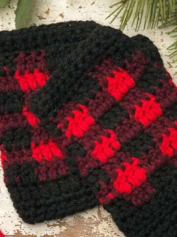 Buffalo Plaid Fingerless Gloves Crochet Pattern featuring red, burgundy, and black plaid stitch fingerless gloves displayed on a rustic wooden background with pine leaf greenery.
