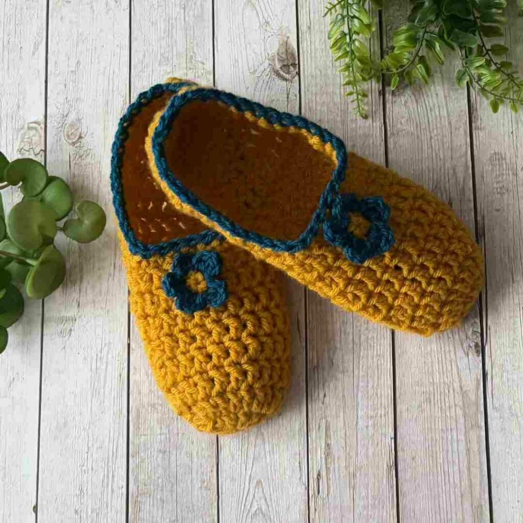 Mustard yellow crochet slippers with blue trim and flower embellishments.