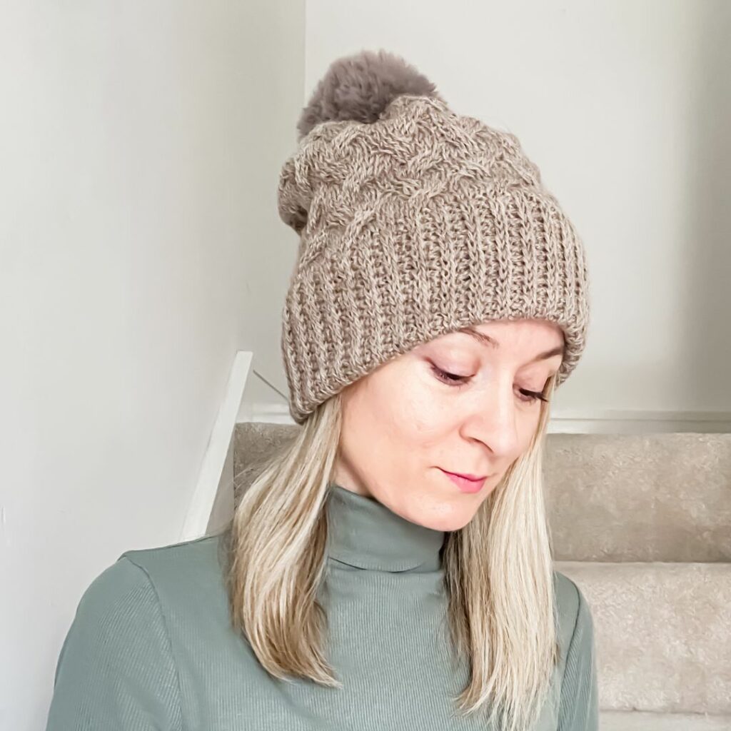 Woman wearing a taupe Tunisian crochet cable hat with a wide ribbed brim and faux fur pom pom.