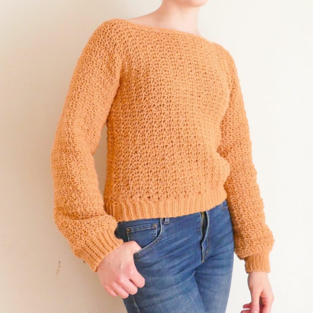 Woman wearing a pumpkin orange textured crochet boatneck sweater with long sleeves and ribbed cuffs and hem.