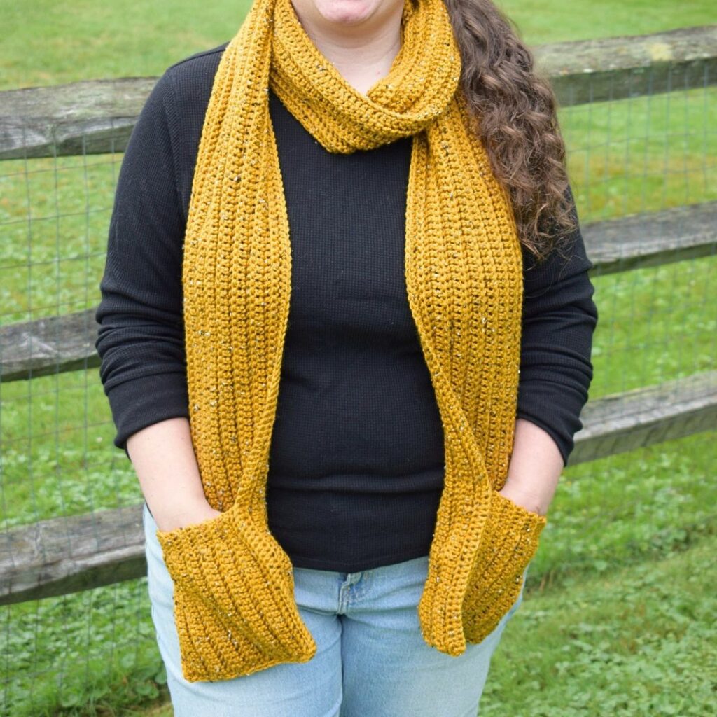 Woman wearing a mustard yellow ribbed crochet scarf with large pockets at each end.