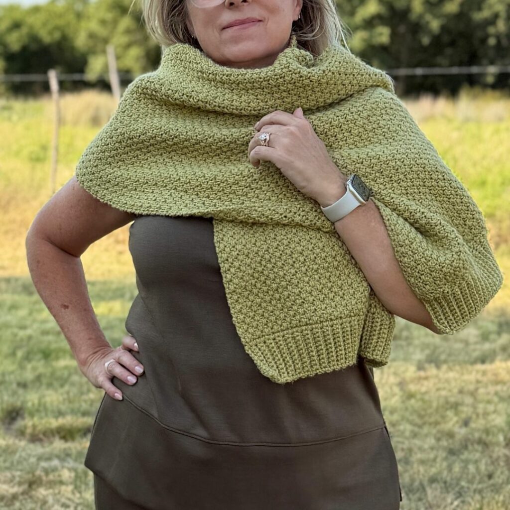 Woman wearing a sage green textured crochet wrap draped around her shoulders with ribbed ends.