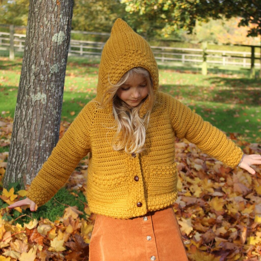 Child wearing a mustard yellow crochet hooded cardigan with long sleeves, textured edging, and front buttons.