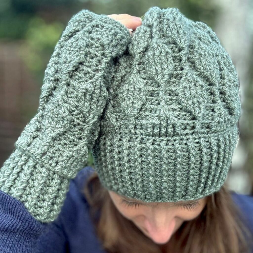 Woman wearing a sage green crochet hat with a ribbed cuff and textured trailing leaf design.