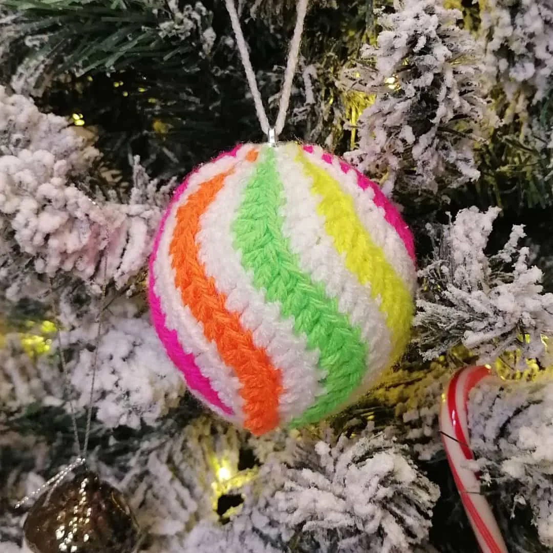 A colorful crochet Christmas bauble ornament hangs on a snow-frosted Christmas tree. The bauble features spiraling stripes in neon green, yellow, orange, hot pink, and white. Behind it, warm fairy lights peek through the branches, and a red-and-white candy cane adds a festive touch.