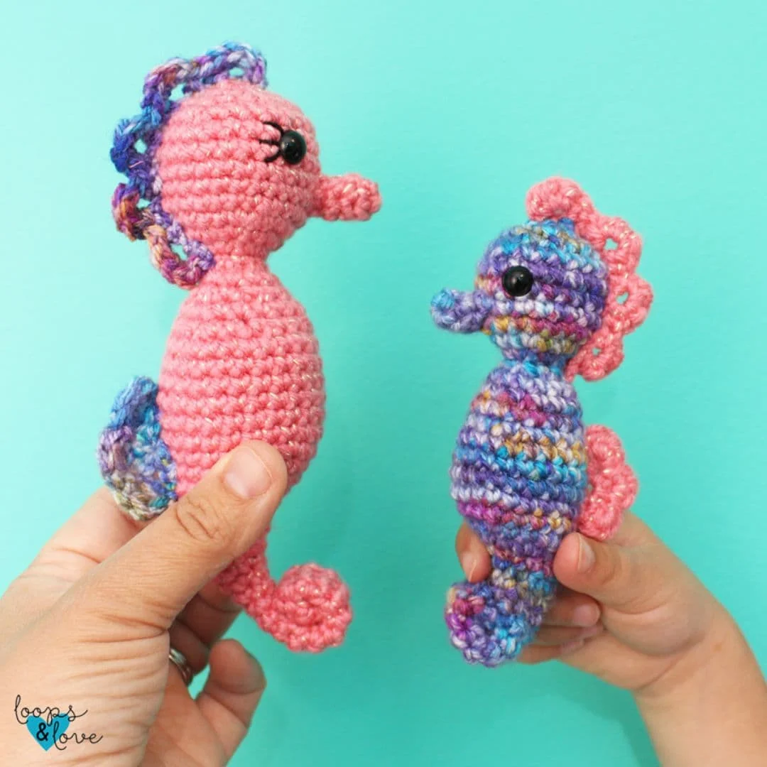 Two crocheted seahorse amigurumi are held up against a turquoise background. The left seahorse is solid pink with a multicolored purple and blue mane and tail, and has embroidered eyelashes. The right seahorse features a multicolored body in blues, purples, and yellows, with pink accents for the fins, tail, and mane. Both have shiny black safety eyes and curled tails.