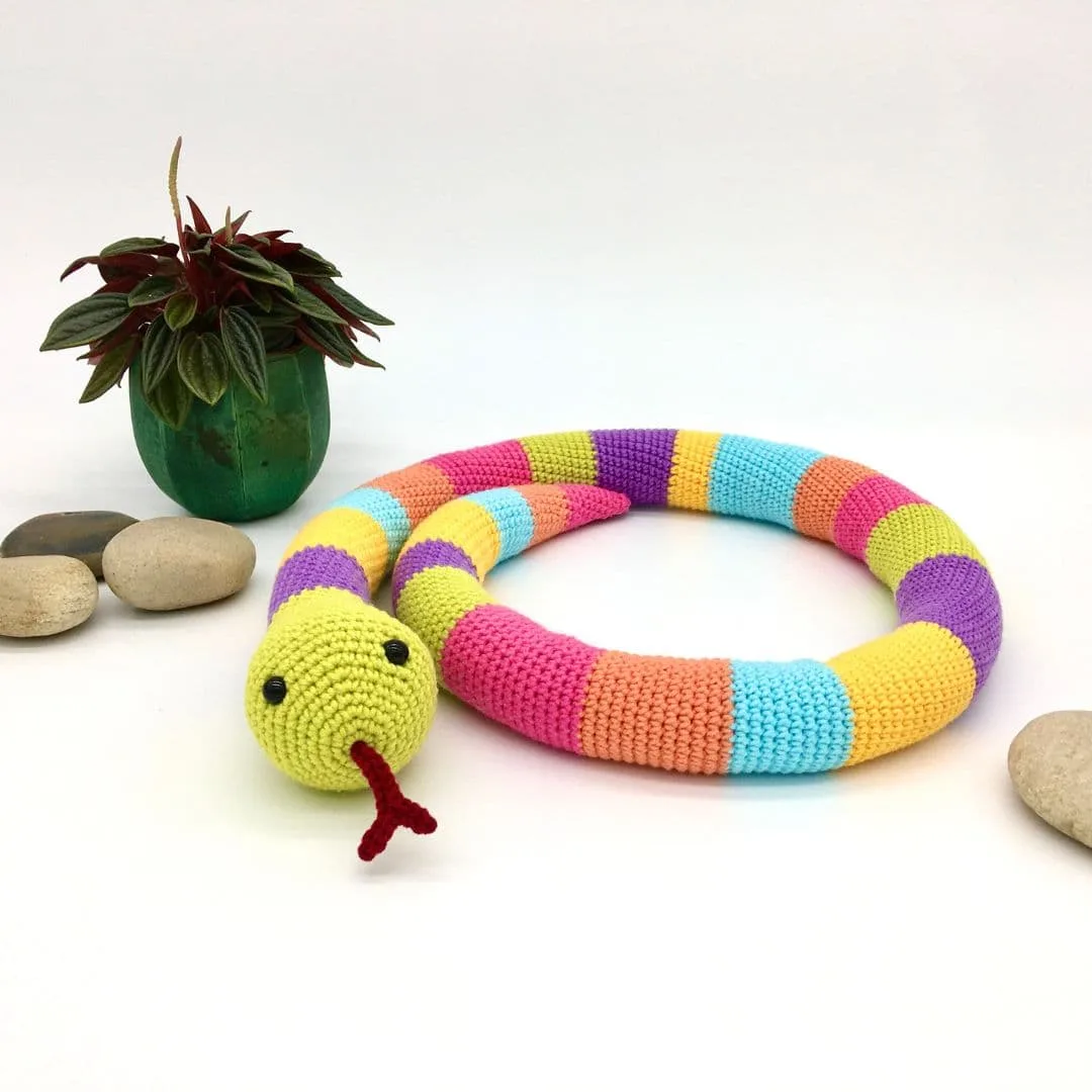 A colorful crocheted snake toy lies coiled on a white surface. The snake is made with alternating bright stripes of lime green, purple, pink, orange, blue, and yellow. It has black safety eyes and a red forked tongue sticking out. To the left is a small green plant in a green pot with red-tipped leaves, and smooth tan stones are scattered nearby.
