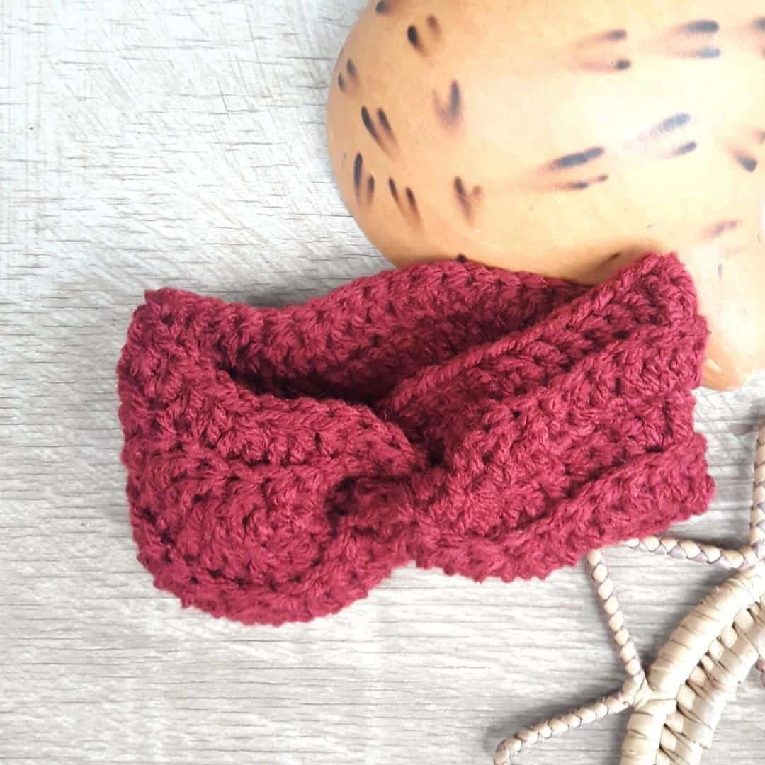 A hand-crocheted headband in a rich burgundy yarn is displayed on a light wood surface. The headband features a twist in the center, creating a stylish knotted effect. Part of a tan gourd with dark markings and a woven raffia decoration are visible in the background, adding natural texture and warmth to the composition.