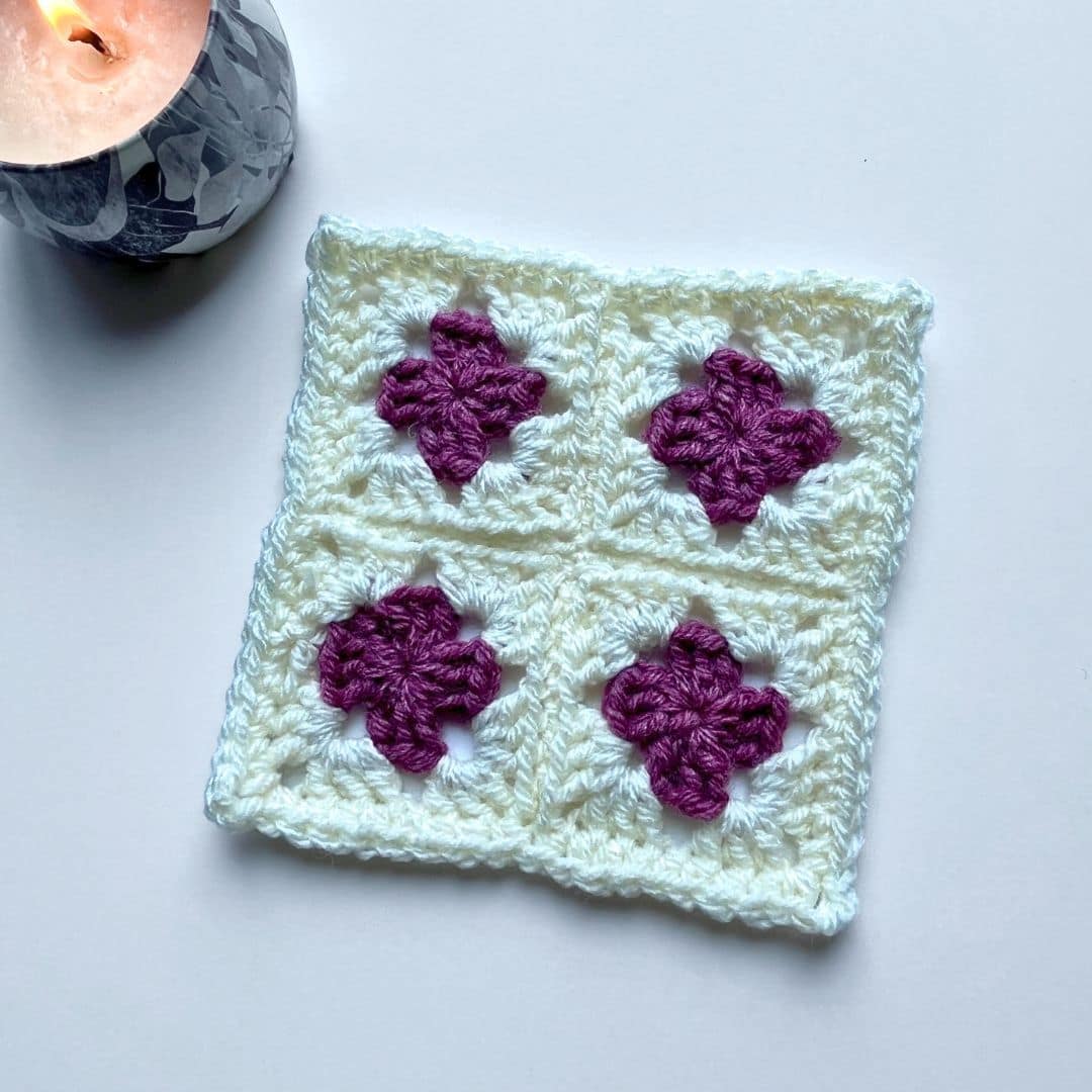 A crochet square made of four smaller granny squares is shown on a white background. Each smaller square features a purple flower-like motif at the center surrounded by a soft white border. In the upper left corner of the image, a marbled gray candle is lit, adding a cozy ambiance to the minimalist and clean setting.