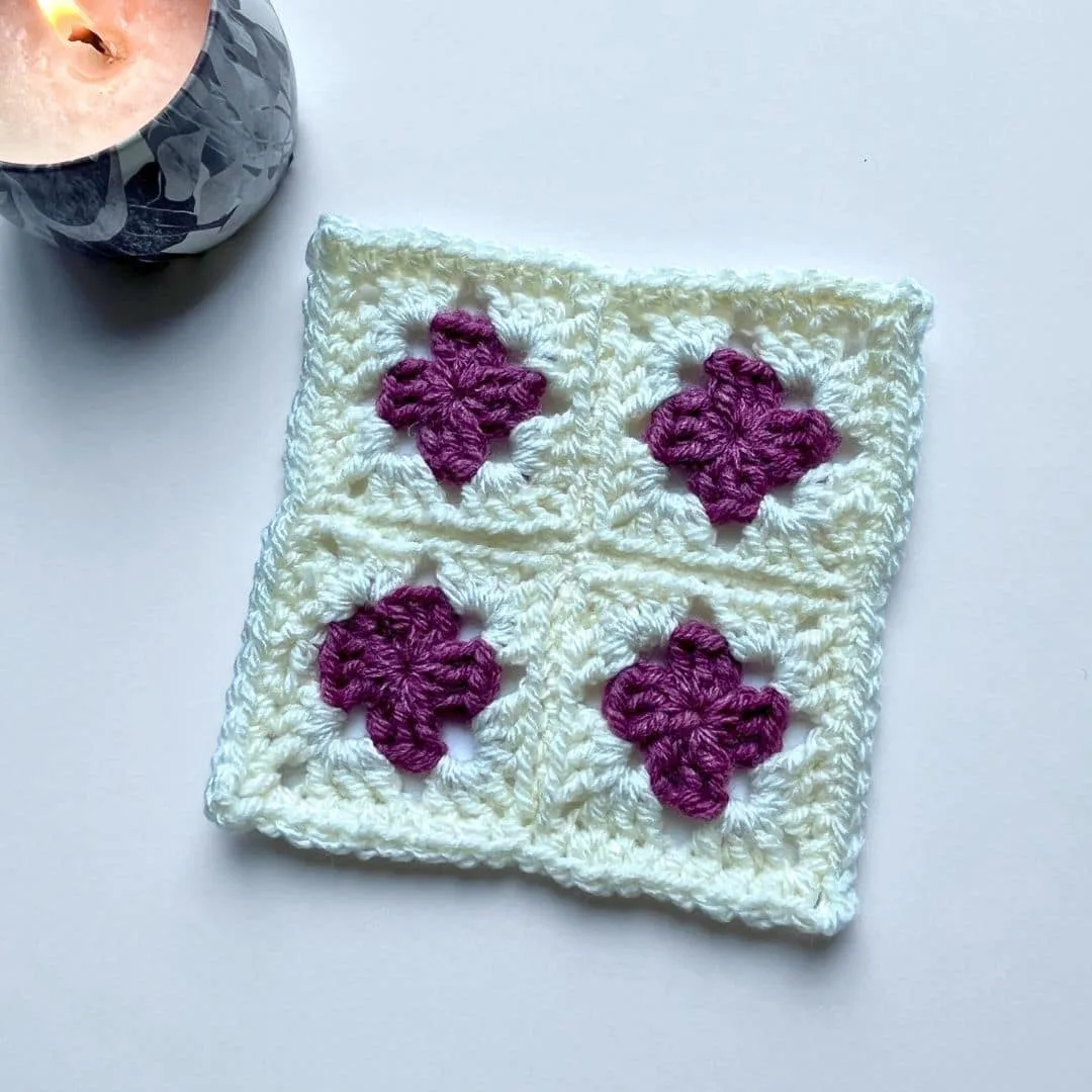 A crochet square made of four smaller granny squares is shown on a white background. Each smaller square features a purple flower-like motif at the center surrounded by a soft white border. In the upper left corner of the image, a marbled gray candle is lit, adding a cozy ambiance to the minimalist and clean setting.