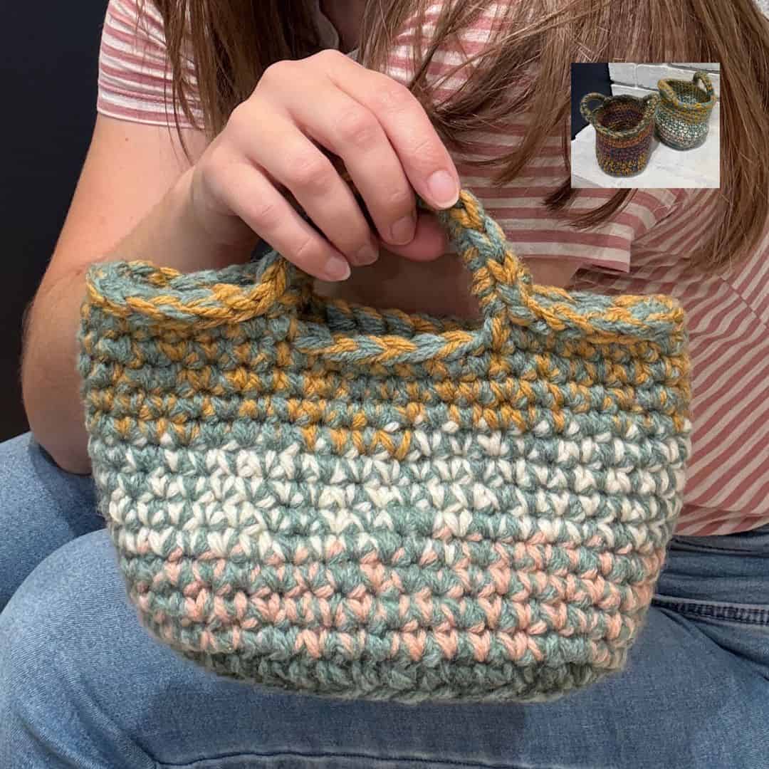 A woman wearing a pink and white striped shirt and jeans holds a small, sturdy crochet basket with built-in handles. The basket features thick, textured stitches in muted pastel shades of sage green, cream, blush pink, and mustard yellow. In the top right corner, a smaller inset image shows two additional crochet baskets in different color combinations placed on a white surface against a dark background.