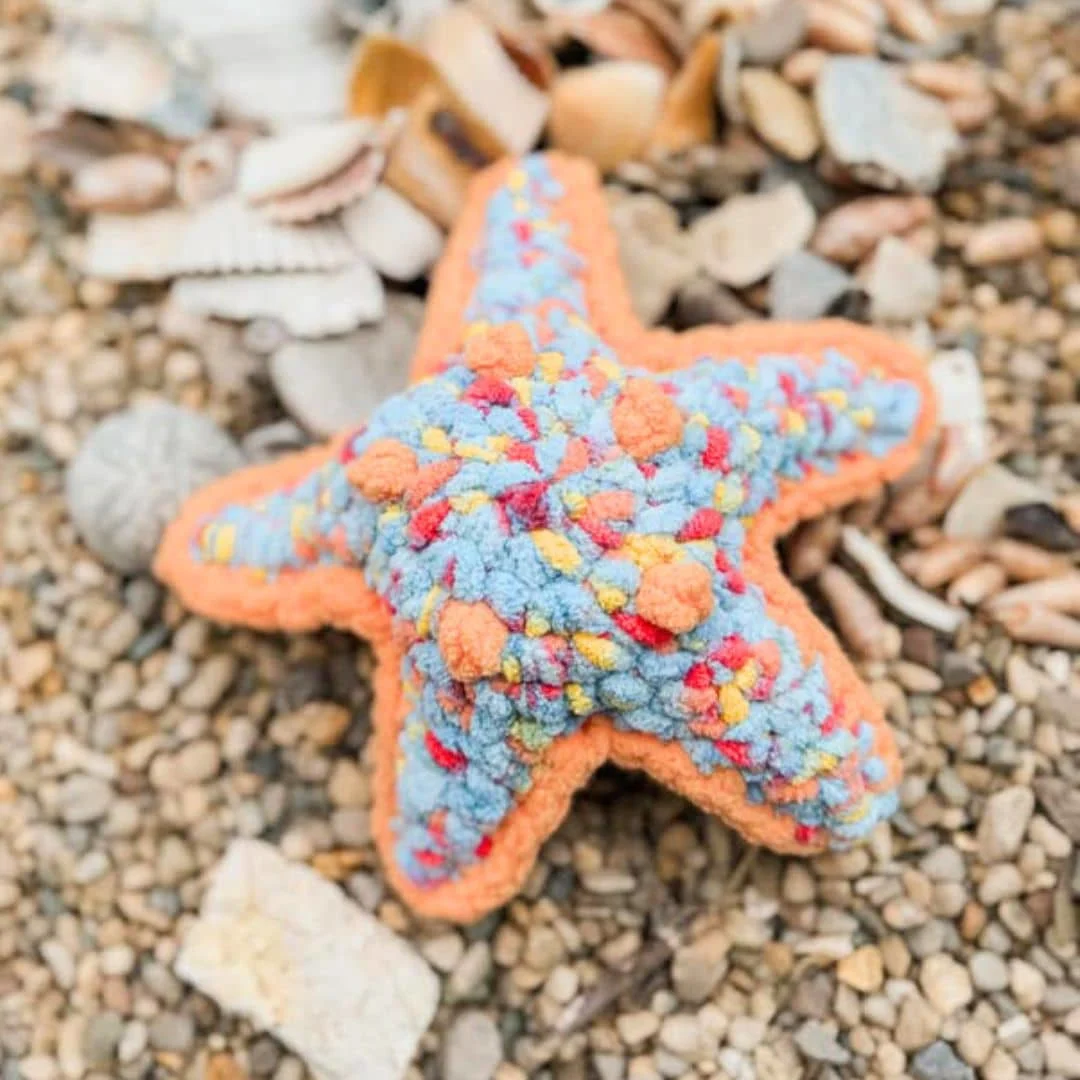 A soft, colorful crochet starfish toy lies on a bed of pebbles and scattered seashells. The starfish is crafted with a vibrant blend of plush blue yarn speckled with red, yellow, and orange, and is outlined with a bold orange border. The texture is bumpy and cozy, giving it a playful, whimsical appearance suitable for children or beach-themed decor.
