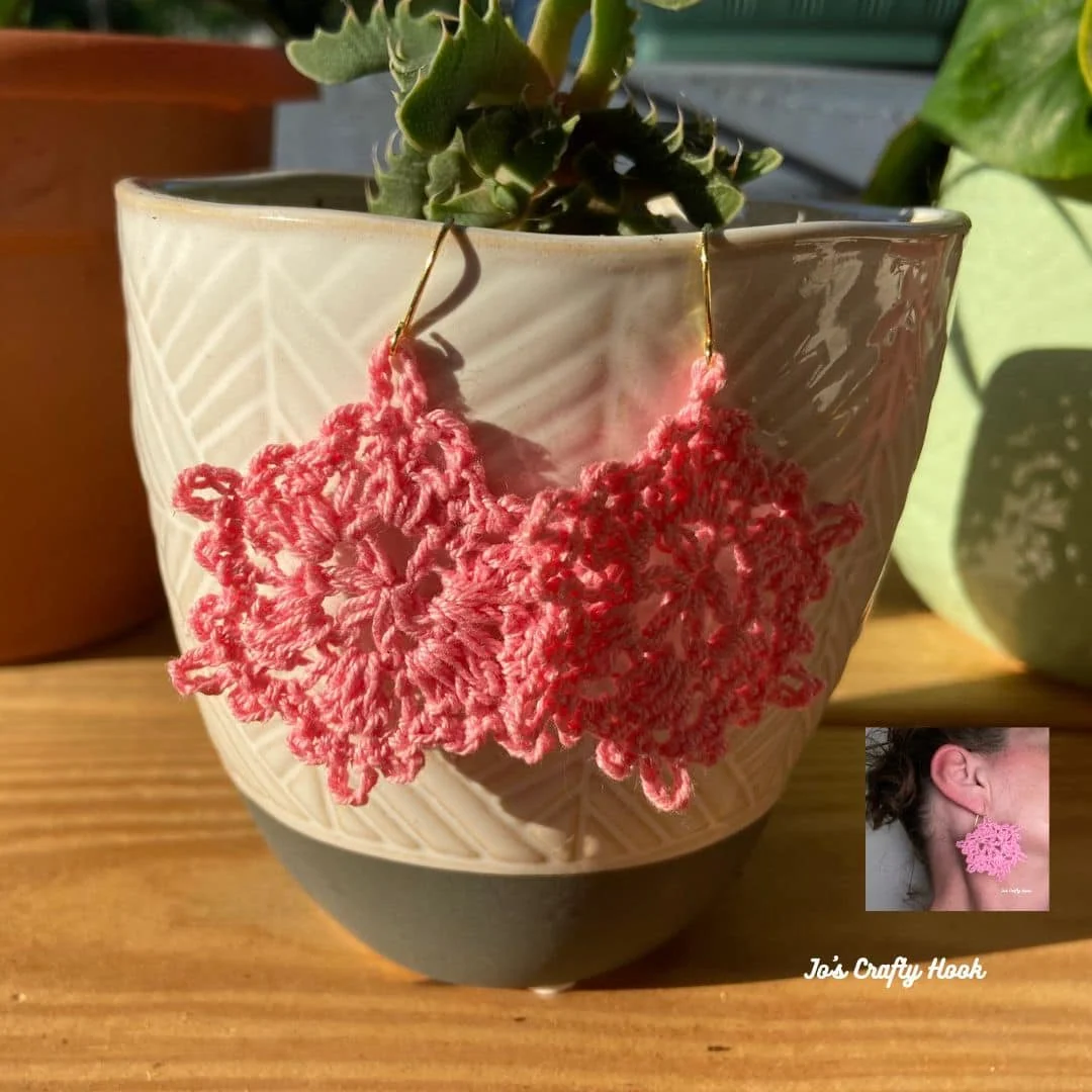 A pair of pink crochet earrings shaped like delicate lace snowflakes hang from the rim of a textured ceramic planter. The earrings are lightweight and intricate, featuring loops and picot points. In the bottom right corner, an inset photo shows one earring worn, highlighting its dainty size and bold yet elegant appearance. The text "Jo's Crafty Hook" is written in white in the corner. The background includes potted succulents and warm natural light on a wooden surface.