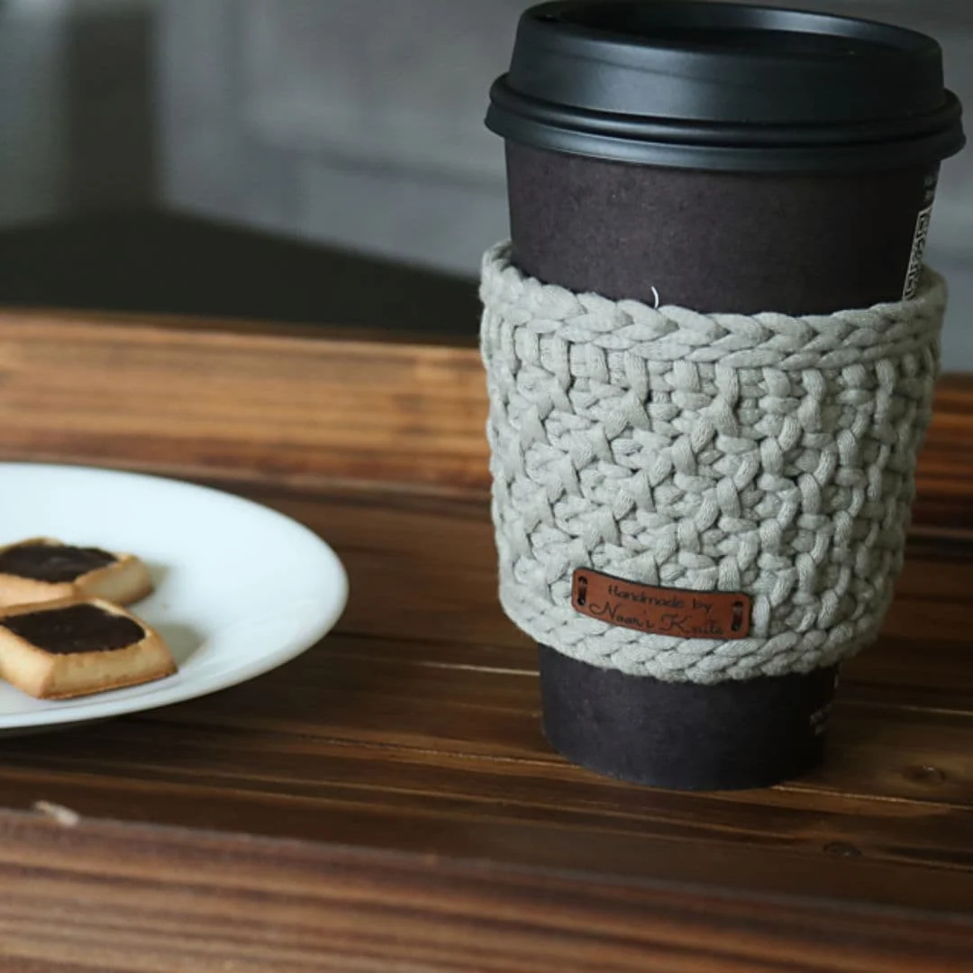 A black takeaway coffee cup is wrapped in a thick, taupe-colored crochet cup cozy made with chunky yarn and a textured stitch. A small faux leather tag on the cozy reads “Handmade by Naomi Knits.” The cup sits on a wooden surface, next to a white plate holding shortbread cookies topped with chocolate. The scene feels warm and cozy, with neutral tones and a handmade charm perfect for gifting or showcasing a functional crochet project.