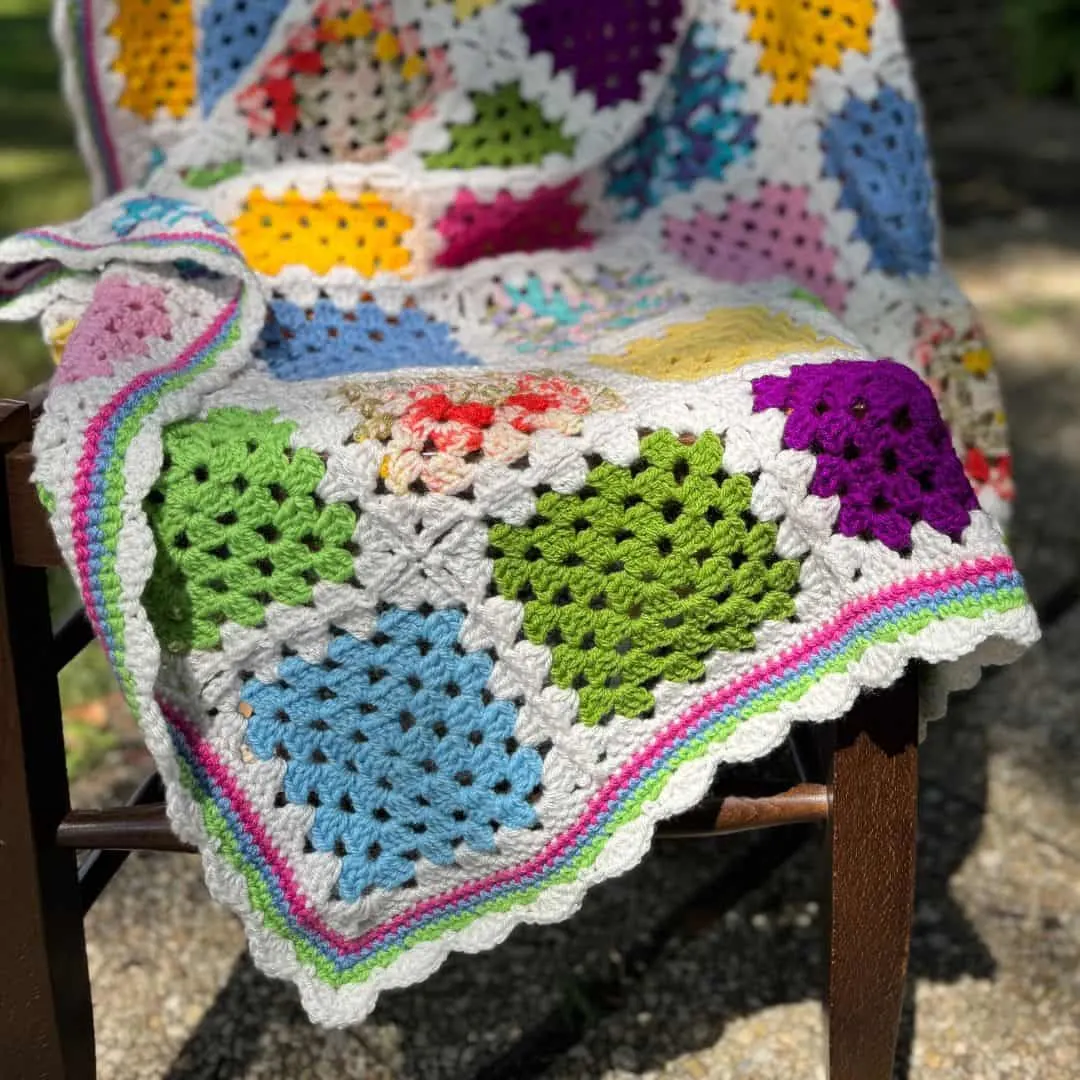 A colorful crochet granny square blanket is draped over a wooden chair outdoors, catching sunlight. The blanket features a cheerful patchwork of granny squares in bright shades of green, blue, pink, purple, yellow, and multicolor prints, bordered in white. A scalloped edging in stripes of green, blue, pink, and white frames the piece. The setting, with dappled light and gravel underfoot, gives the scene a cozy, handmade feel—perfect for showcasing a vibrant, classic crochet project.