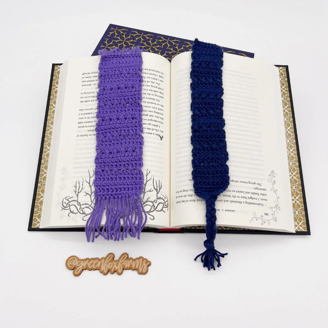 Two crocheted bookmarks—one purple with fringe and one navy with a twisted tassel—rest on the open pages of a hardcover book. A wooden tag reading "@greenfoxfarms" is placed below the book on a white background.
