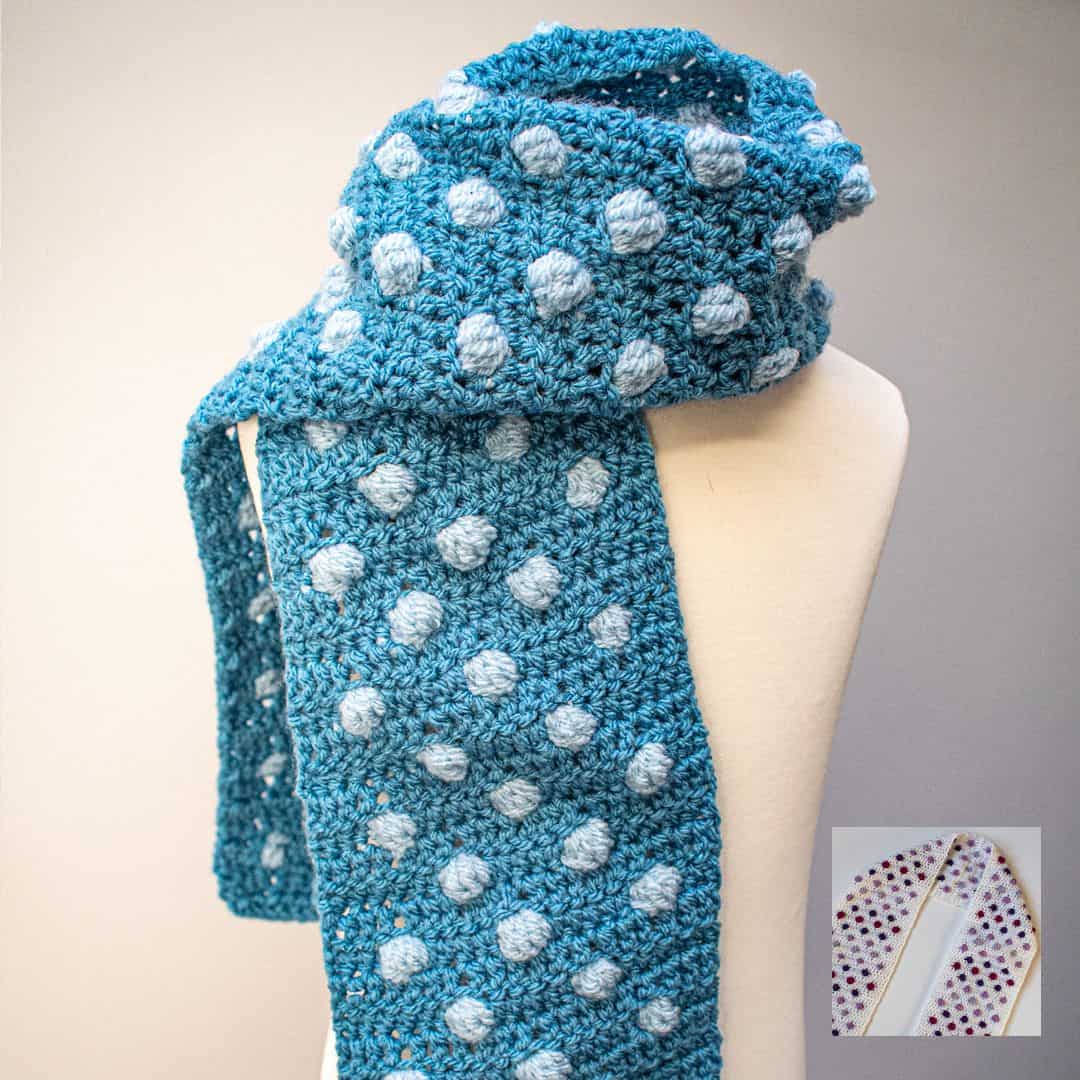 A blue crochet scarf with raised light blue bobble dots is wrapped around a cream-colored mannequin bust. The scarf features a repeating pattern of textured polka dots, giving it a playful and cozy appearance. The yarn has a soft, slightly heathered texture. In the lower right corner, a small inset image shows a different version of the scarf in cream with purple and red bobbles. The background is a soft gray, creating a clean, minimalist look that highlights the stitch detail and texture of the scarf.