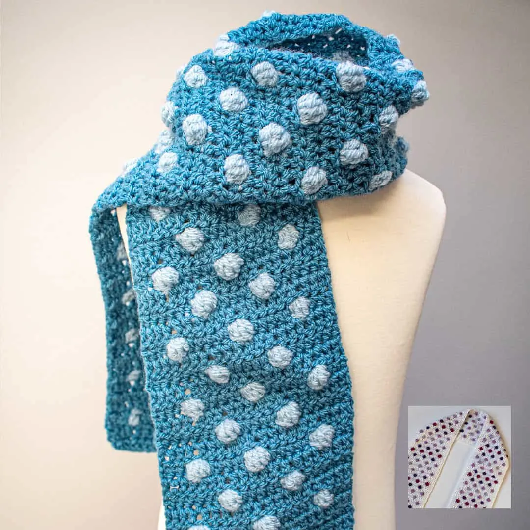 A blue crochet scarf with raised light blue bobble dots is wrapped around a cream-colored mannequin bust. The scarf features a repeating pattern of textured polka dots, giving it a playful and cozy appearance. The yarn has a soft, slightly heathered texture. In the lower right corner, a small inset image shows a different version of the scarf in cream with purple and red bobbles. The background is a soft gray, creating a clean, minimalist look that highlights the stitch detail and texture of the scarf.