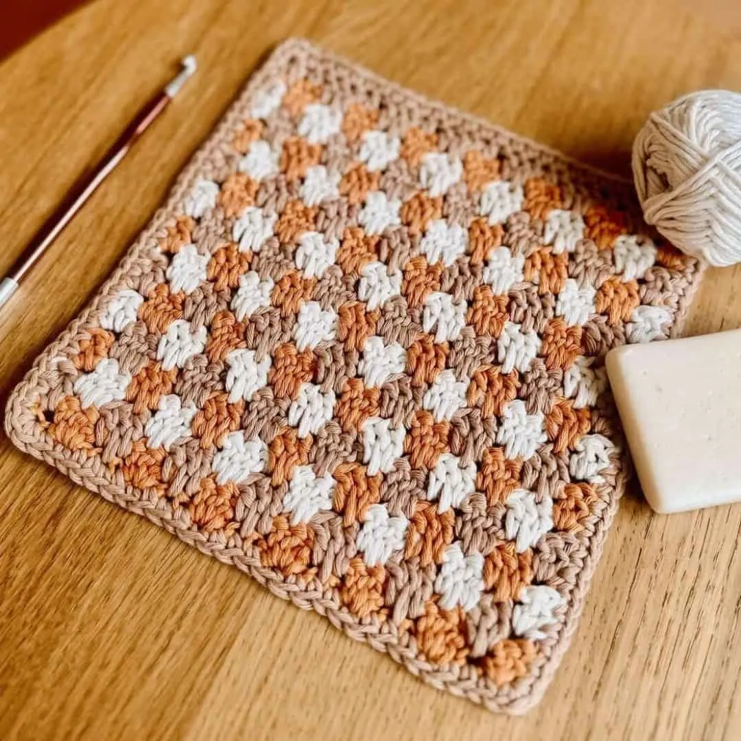 cream, tan, and burnt orange. The stitches are tightly worked in rows of alternating colors, creating a textured, woven appearance. To the right of the cloth is a ball of cream yarn and a natural bar of soap, while a crochet hook with a white handle lies to the left. The cozy, rustic palette and practical materials suggest a handmade item perfect for kitchen or bath use.