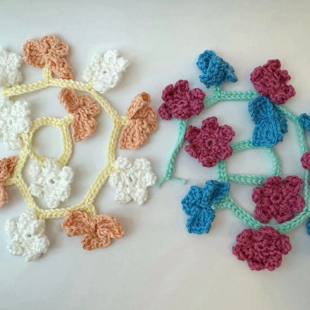 Two delicate crochet flower garlands are displayed on a light surface. The garland on the left features soft white and peach-colored five-petal flowers connected by a pale yellow chain in a circular, web-like design. The garland on the right displays bright fuchsia and rich blue flowers with more layered petals, connected by a light teal chain resembling stems. The contrast between the pastel and vibrant colors adds visual interest, showcasing two different styles of crocheted floral motifs.