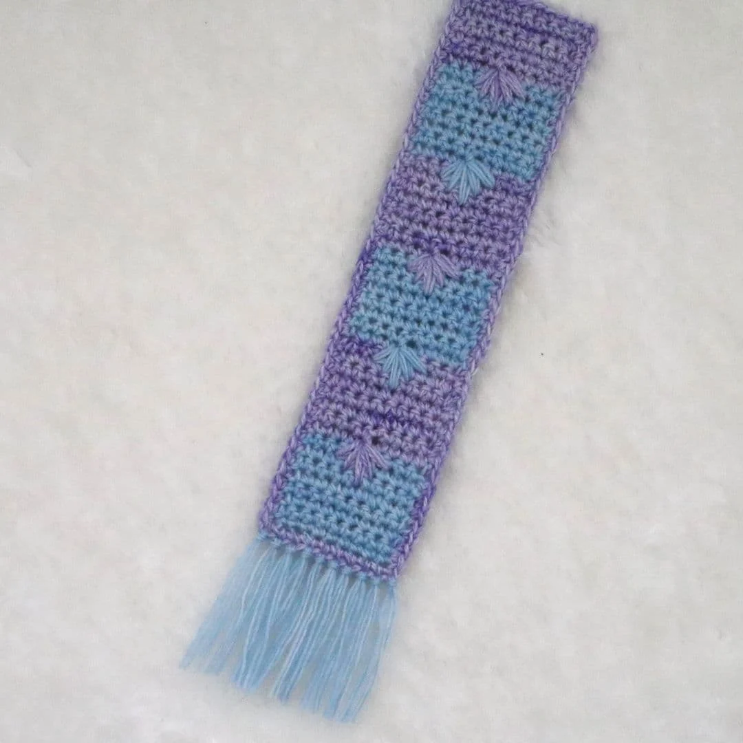 A handmade crochet bookmark in shades of lavender and sky blue rests on a soft white surface. The rectangular design features alternating blocks of color with a starburst stitch pattern at each color transition, creating a subtle textured effect. The bottom is finished with light blue fringe, giving the piece a delicate, decorative touch.