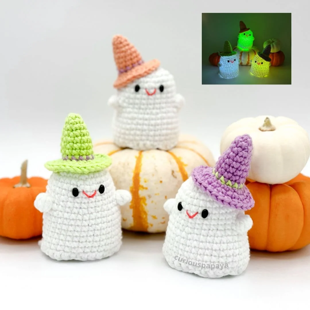 Three crocheted ghost plushies are arranged in front of mini pumpkins on a white background. Each ghost has a cute smiling face and wears a colorful witch hat—one in peach, one in lime green, and one in purple. A small inset photo in the upper right corner shows the same ghosts glowing in the dark with LED lights inside, casting green, yellow, and blue hues. The ghosts are handmade and whimsical, perfect for Halloween decor.