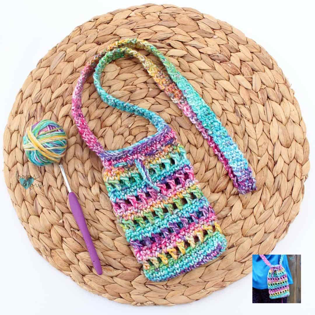 A colorful crocheted crossbody bag lies on a round woven placemat. The bag features a vibrant rainbow yarn with open stitchwork details and a long strap. A matching multicolor yarn ball and a purple-handled crochet hook rest beside it. In the bottom-right corner, an inset image shows the bag being worn over the shoulder with a bright blue shirt, showcasing its size and fit.
