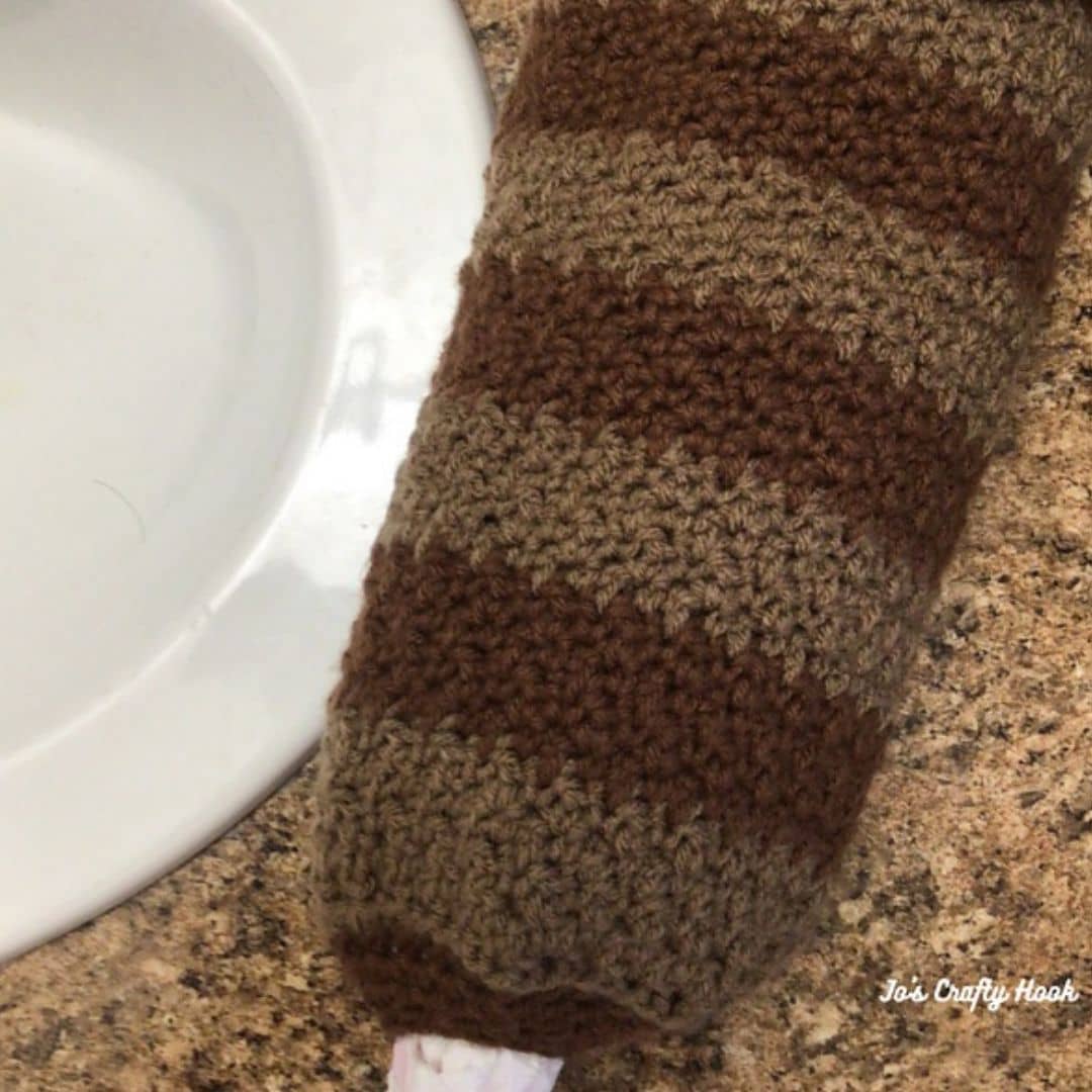 A striped crochet plastic bag holder in shades of brown hangs over a kitchen or bathroom countertop. The textured stitchwork creates alternating bands of medium and dark brown. A white plastic bag peeks out from the bottom opening of the holder. A white sink and part of the countertop are visible in the background. The bottom right corner includes the watermark "Jo’s Crafty Hook."