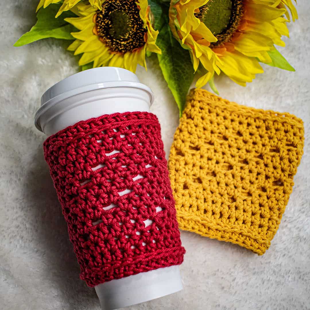 A white travel coffee cup is wrapped in a vibrant red crochet cozy featuring an openwork stitch pattern. Beside it lies a matching yellow crochet cozy with a similar texture. Both items are displayed on a soft, cream-colored fabric surface with three large sunflowers in the background, adding a cheerful, cozy feel to the image.