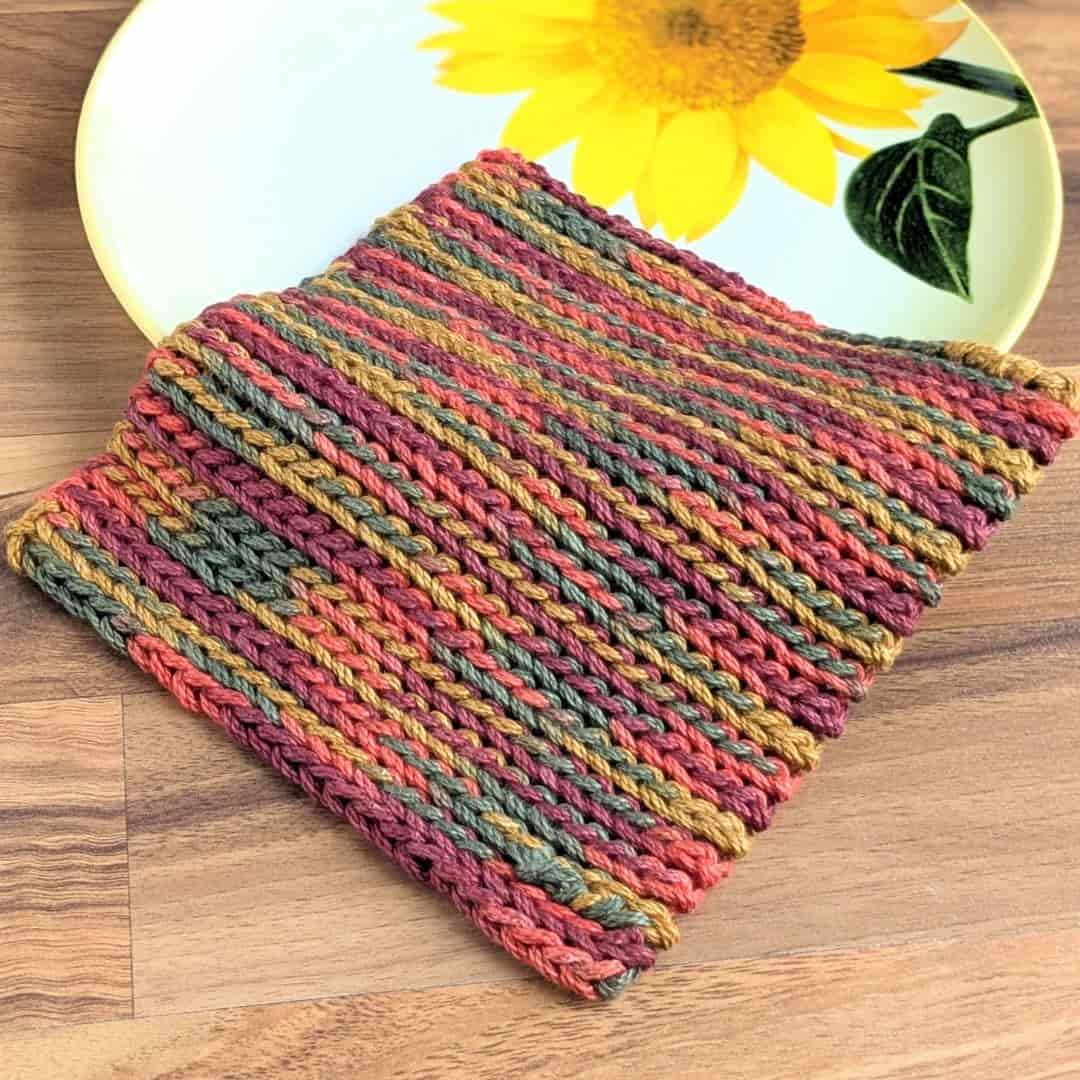 A ribbed crochet dishcloth made with variegated yarn in autumnal shades of olive green, rust, gold, and burgundy is laid flat on a wooden surface. The textured stitches run diagonally across the square. Partially beneath the dishcloth is a decorative plate featuring a large yellow sunflower with green leaves, adding a cheerful contrast to the earthy tones of the yarn.