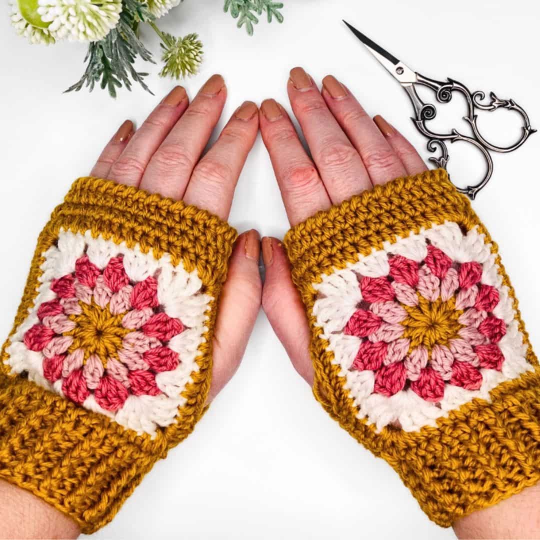 A pair of hands wearing crochet granny square wrist warmers in autumn tones. The wrist warmers feature a central sunburst granny square in mustard, blush pink, and burgundy, bordered in cream, with a solid mustard yellow ribbed cuff and trim. The hands are placed flat on a white surface, surrounded by small faux greenery, flowers, and a pair of ornate black embroidery scissors. The overall aesthetic is clean and cozy with a vintage touch.