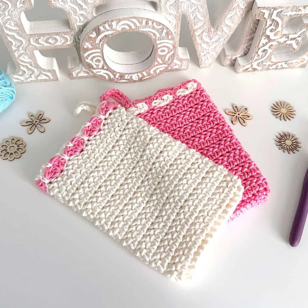 Two crocheted rectangular pouches with decorative heart details at the top edge—one in solid cream with pink accents and the other in solid pink with cream accents—displayed on a white surface with wooden flower embellishments and a carved “HOME” sign in the background.