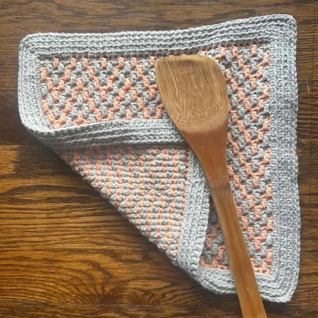 Crocheted potholder featuring a mosaic-style pattern in peach and gray yarn, bordered with solid gray, and folded to reveal the reverse side. A wooden spatula rests on top, with the piece displayed on a wooden surface.