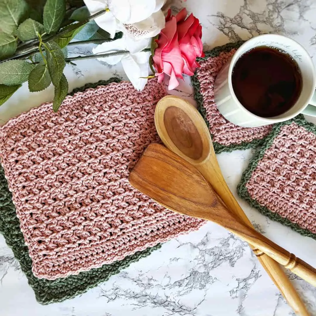 Crocheted dishcloths and coasters in a textured dusty rose yarn with sage green trim are displayed on a marble surface. A wooden spoon and spatula are placed across the dishcloth, with a green mug of coffee and pink and white roses adding a cozy and elegant kitchen touch.