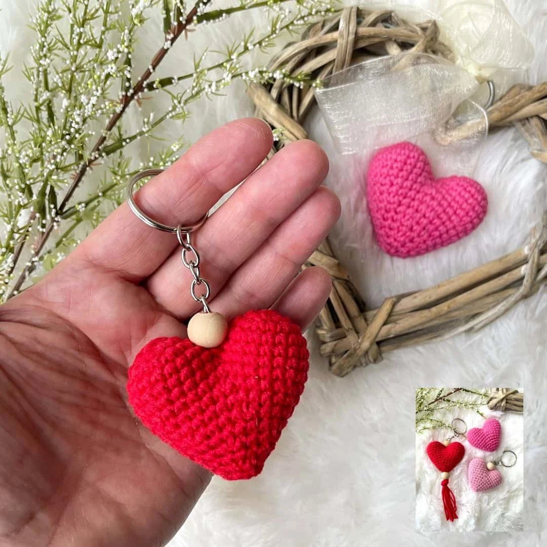 Crocheted heart keychain in bright red yarn with a wooden bead, held in a person's hand. In the background, a pink crocheted heart is displayed in a rustic wooden heart frame, and a small inset image shows three crocheted heart keychains in red, dark pink, and light pink on a soft white surface.