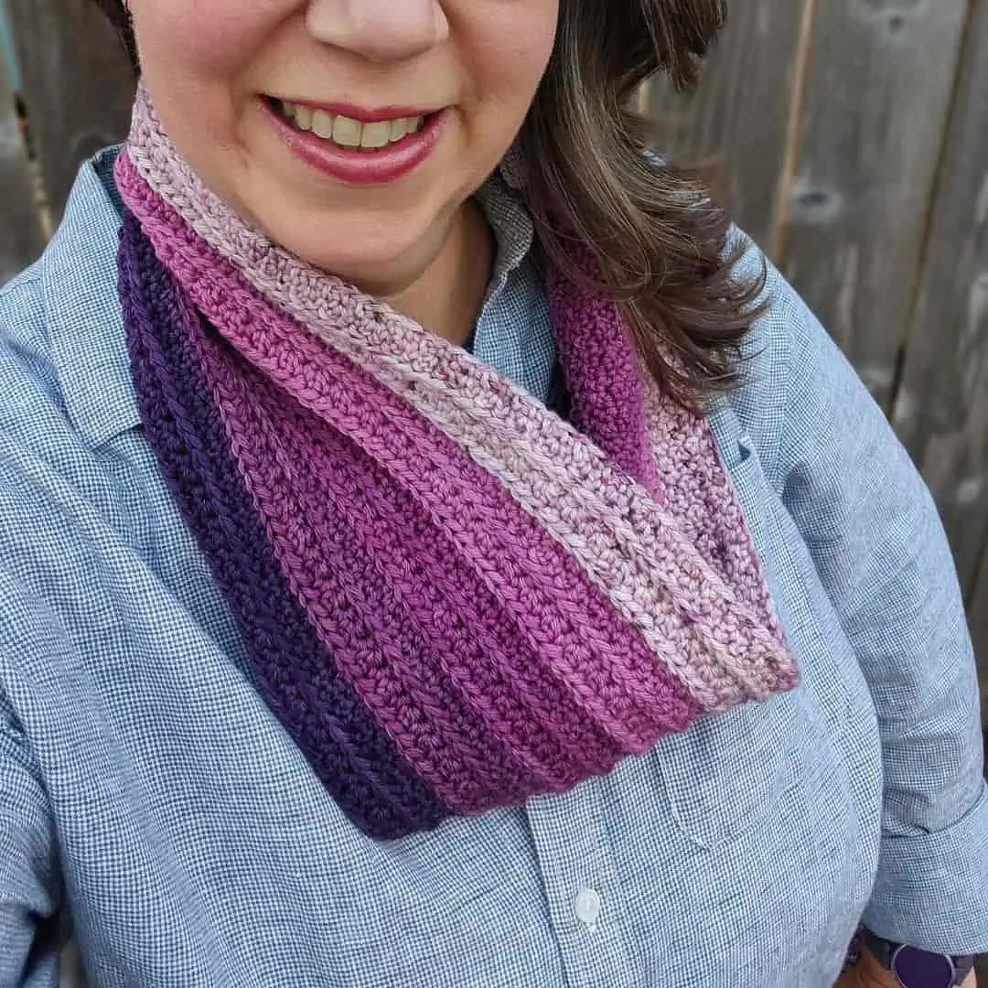 A woman wearing a striped crochet infinity scarf in shades of purple, plum, and lavender. The textured ribbing highlights the color gradient. She is dressed in a light blue checkered shirt, smiling in front of a wooden fence.