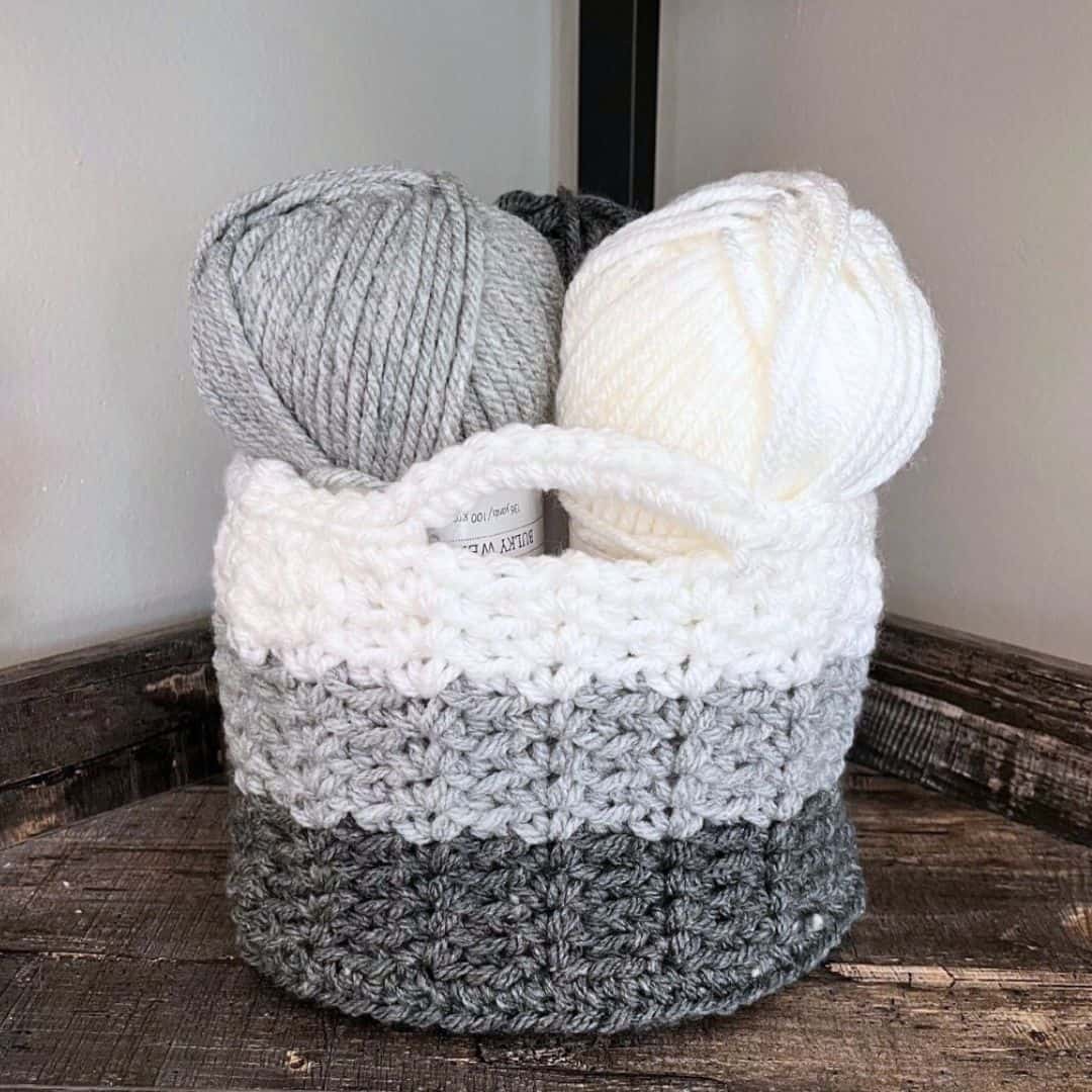 A crochet storage basket with sturdy handles sits on a rustic wooden surface in the corner of a room. The basket features three color blocks in dark gray, medium gray, and white, and is filled with matching yarn skeins in white, gray, and charcoal.