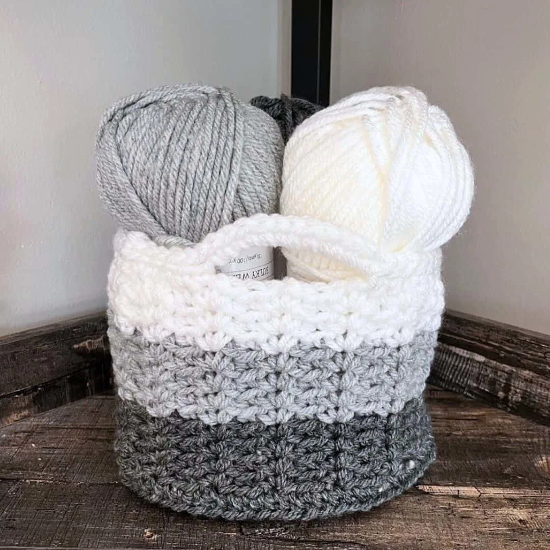 A crochet storage basket with sturdy handles sits on a rustic wooden surface in the corner of a room. The basket features three color blocks in dark gray, medium gray, and white, and is filled with matching yarn skeins in white, gray, and charcoal.