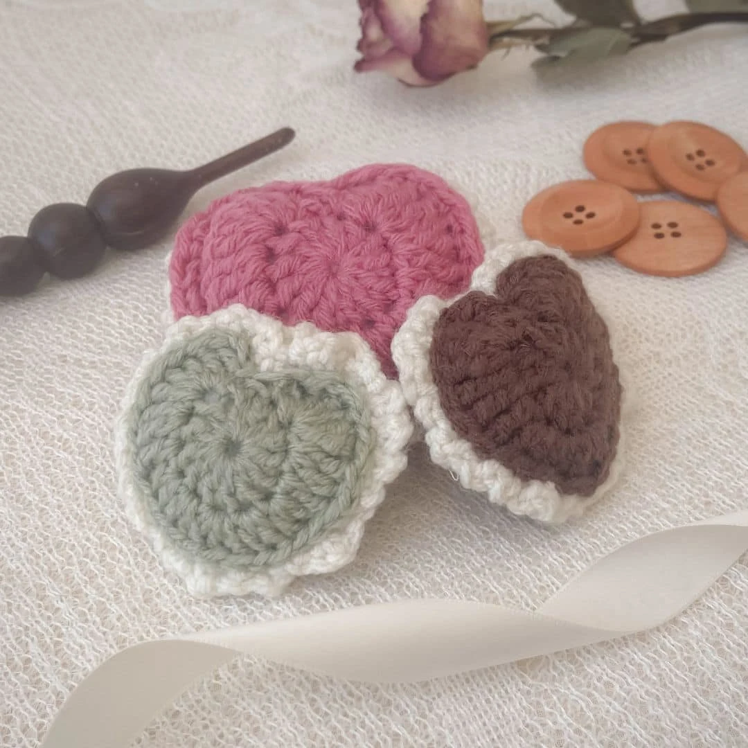 Three crochet heart-shaped macarons are displayed on a soft lace surface. The macarons come in shades of rose pink, mint green with a cream border, and chocolate brown with a cream border. Surrounding them are wooden buttons, a cream ribbon, a crochet hook with a beaded handle, and a dried rose for a cozy, vintage aesthetic.