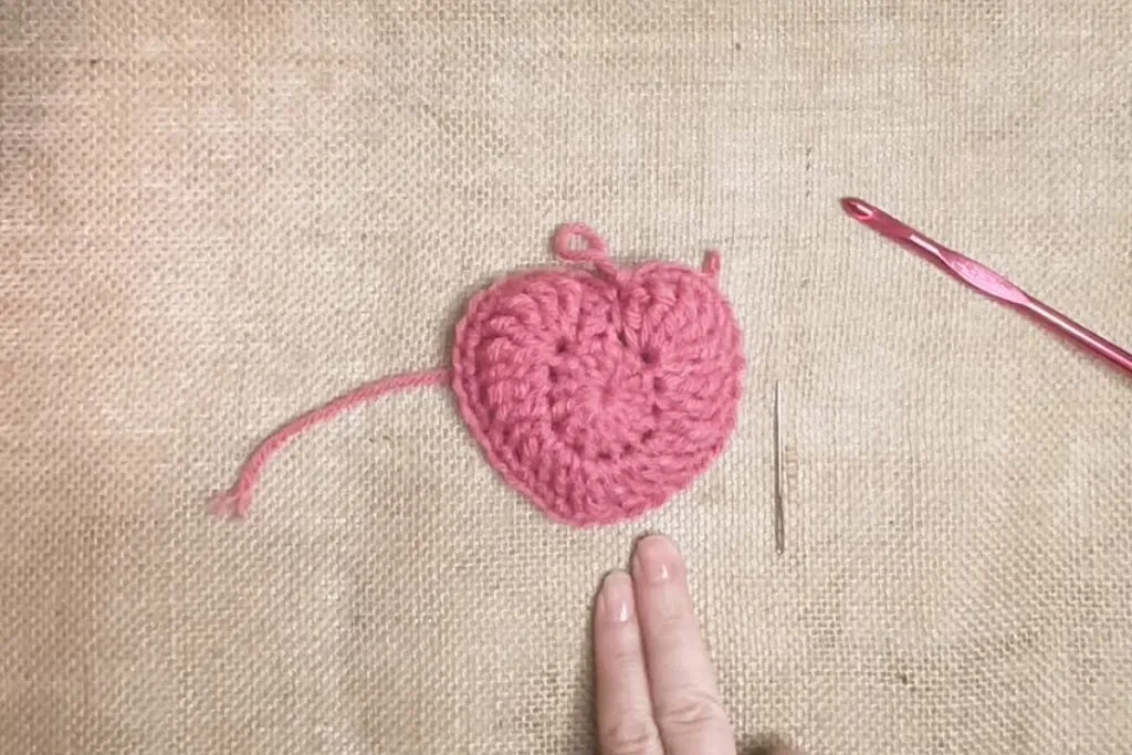 Close-up of a pink crochet heart motif in progress, shown on a burlap background with a red crochet hook and a tapestry needle nearby. Two fingers at the bottom of the image provide scale.