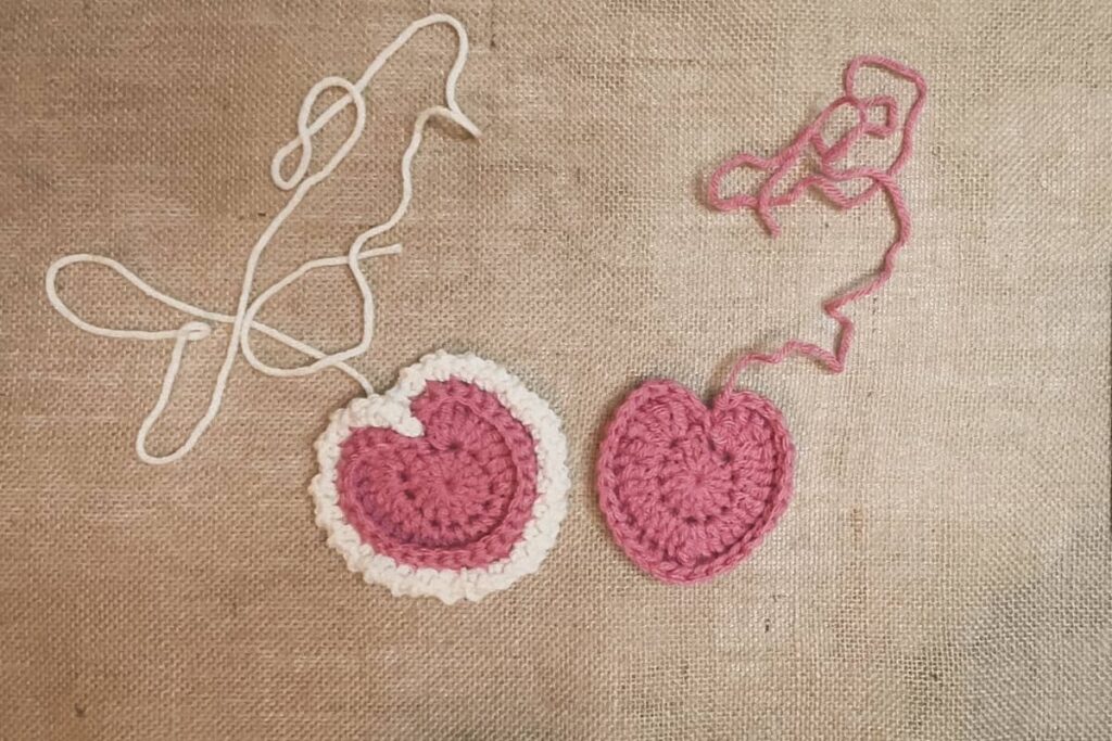 Two pink crochet heart motifs on a burlap surface, one edged with white scalloped stitches and the other plain. Both hearts have yarn tails still attached, indicating work in progress.