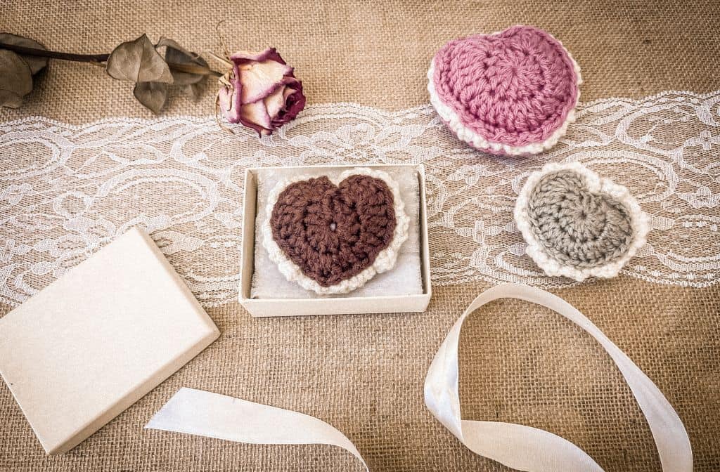 Flat lay of handmade crochet puffed heart keepsakes in pink, brown, and sage green with lace edging. The brown heart is displayed in a small gift box, surrounded by a dried rose, ribbon, and decorative lace on a burlap background—perfect for gifting or sentimental keepsakes.