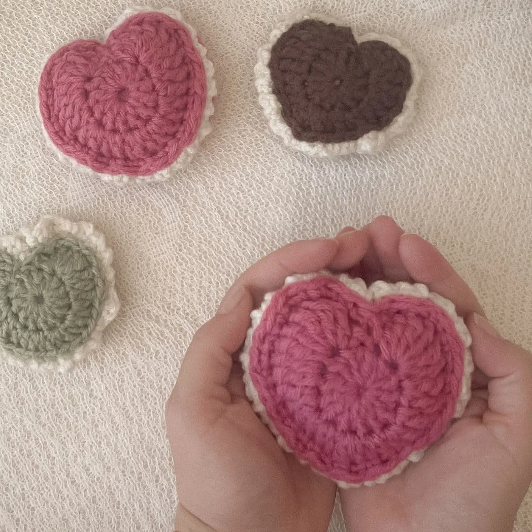 A pink puffed crochet heart keepsake held in a hand, with additional hearts displayed on a soft background to show scale and size options.